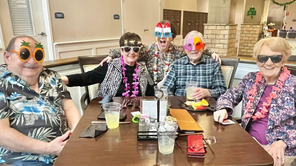 Smiling seniors, sporting sunglasses and leis, savoring a delightful happy hour at their senior living residence, while a staff member captures the joyous occasion.