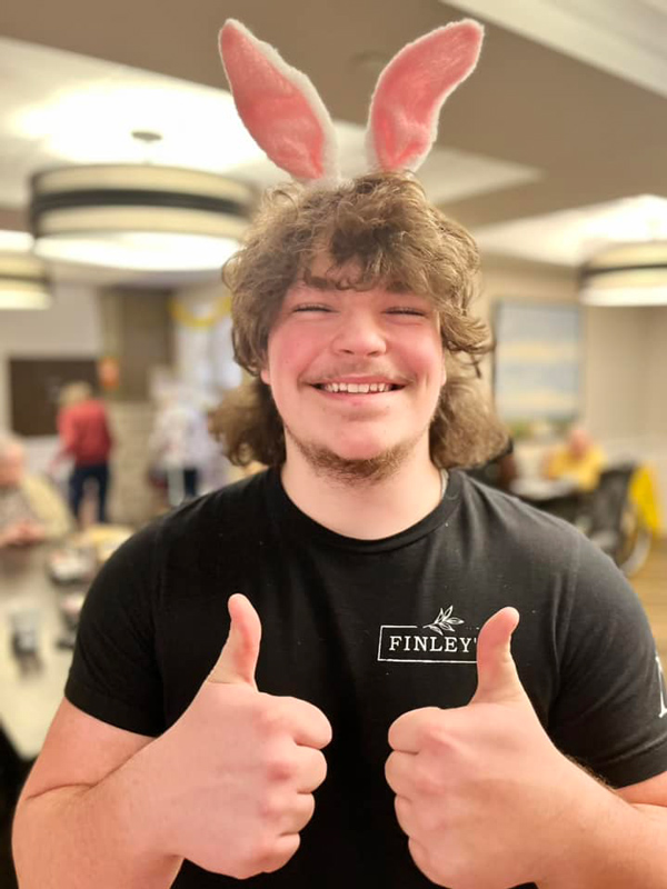 A smiling young man wearing pink bunny ears gives two thumbs up, celebrating Happy Easter in a cheerful atmosphere at a senior living community.