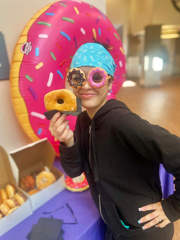 A smiling person wearing fun donut-shaped glasses and a blue bandana playfully holds up a donut in celebration of National Donut Day, standing in front of a large inflatable donut.