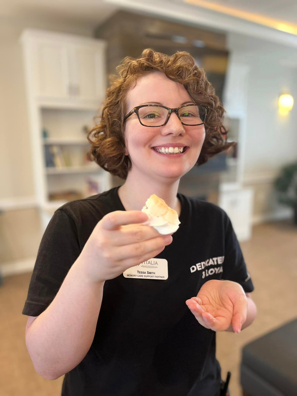 A smiling woman in a black 'DEDICATED & LOYAL' shirt enjoys a treat during the US News Award celebration at Vitalia Senior Living, holding a small dessert in her hand.