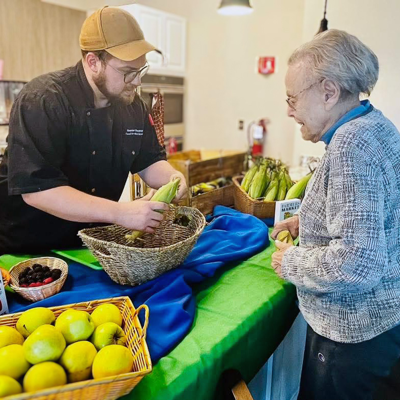 Team member helps a senior resident select fresh corn at a farmers market–style table filled with apples, berries, and produce baskets in the community.