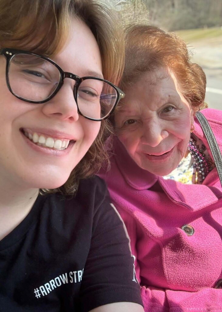 A young woman takes a cheerful selfie with a senior resident in a bright pink coat, both smiling warmly while enjoying time together in the community.