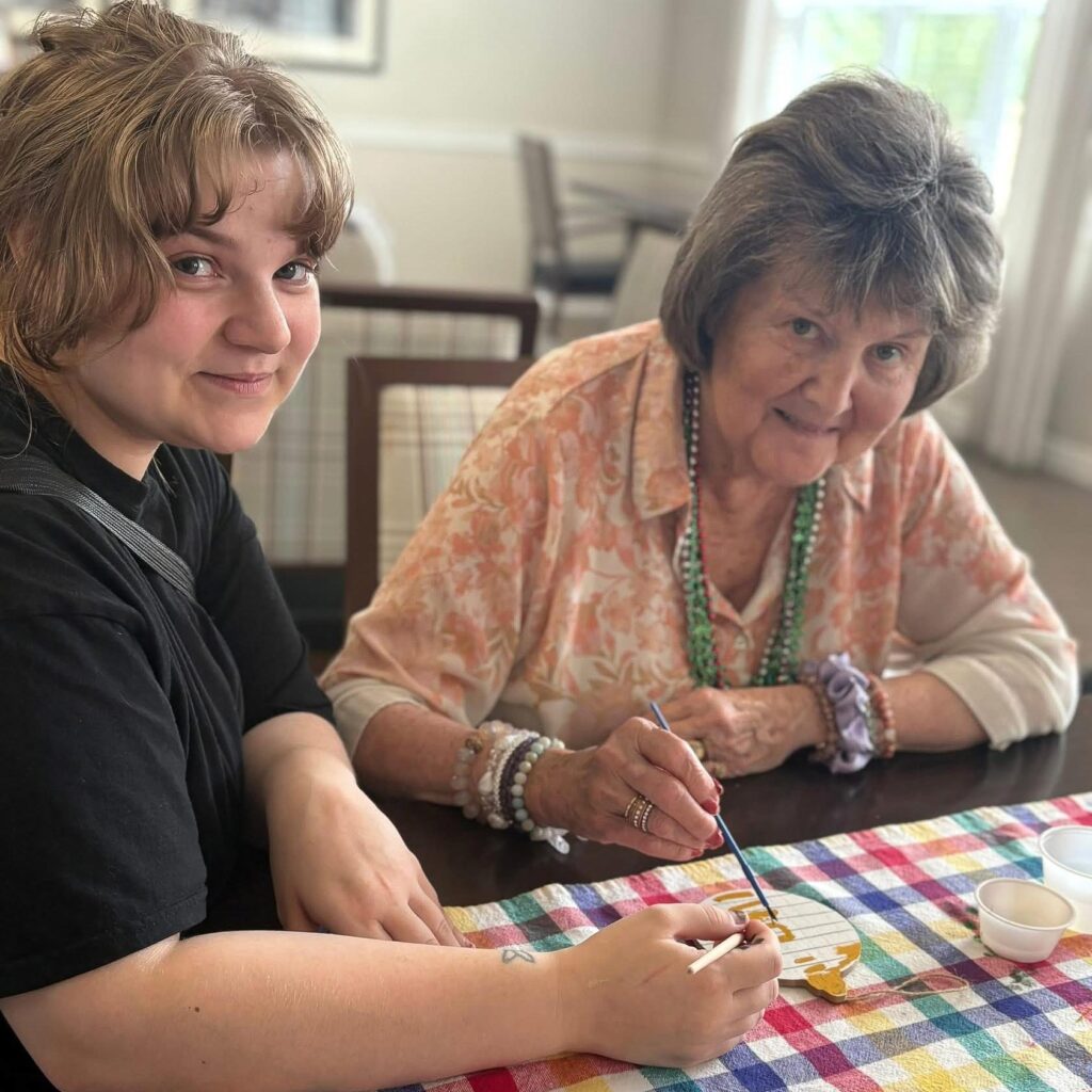 A senior resident and a young woman sit together at a table, smiling while painting on a wooden craft project during a creative activity in the community.