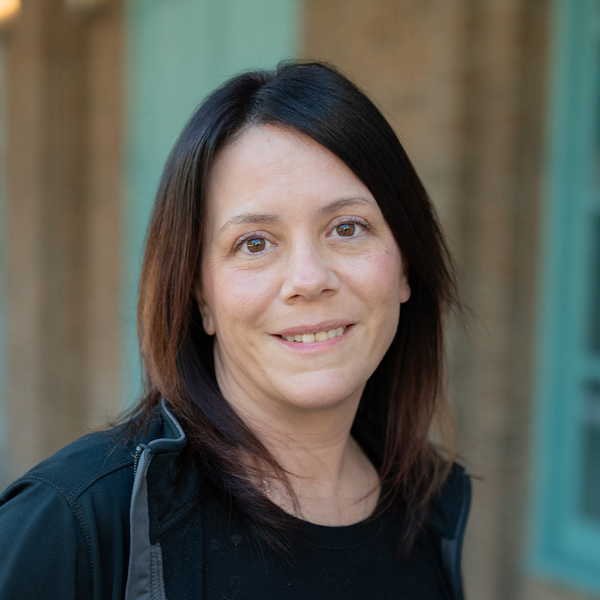Brandy Morris, Wellness Director at Vitalia Strongsville, in a professional outdoor headshot with straight dark shoulder-length hair and softly blurred brick and teal window details in the background.