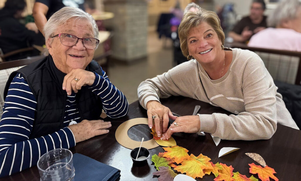 A resident at Vitalia Strongsville smiles alongside a loved one, enjoying a crafting session in an elegant common space.
