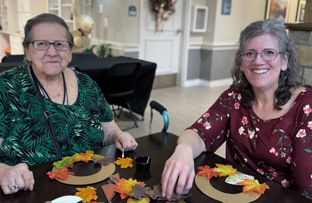 A resident and loved one smile during a relaxed crafting session, making fall wreaths.