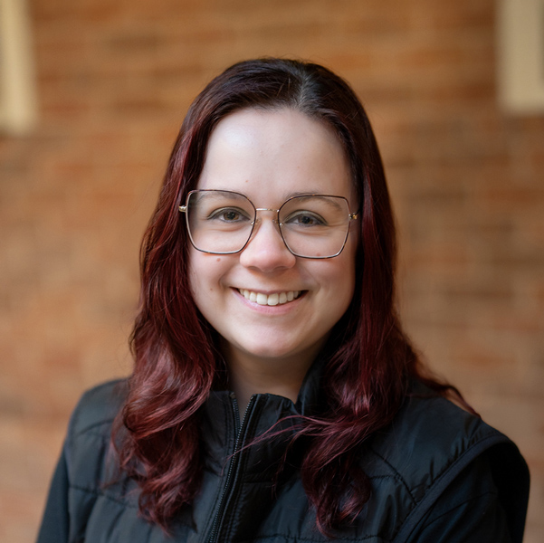 Bethany Sara, Business Office Director at Vitalia Strongsville, smiling in a professional outdoor headshot with long dark red hair, large glasses, a black vest, and a softly blurred brick background.