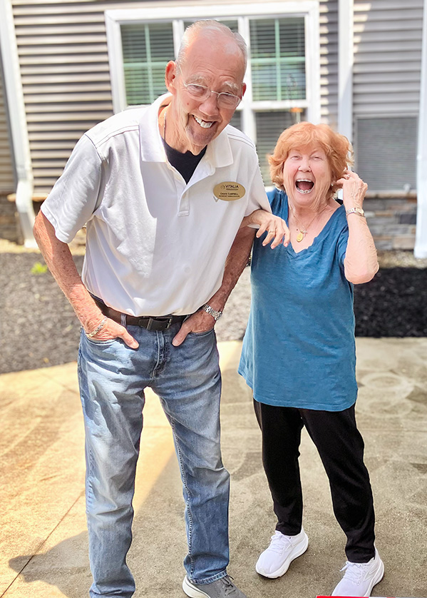 A senior man wearing a gold Resident Ambassador badge smiles with a senior woman, her face lit up in laughter, at Vitalia Strongsville.