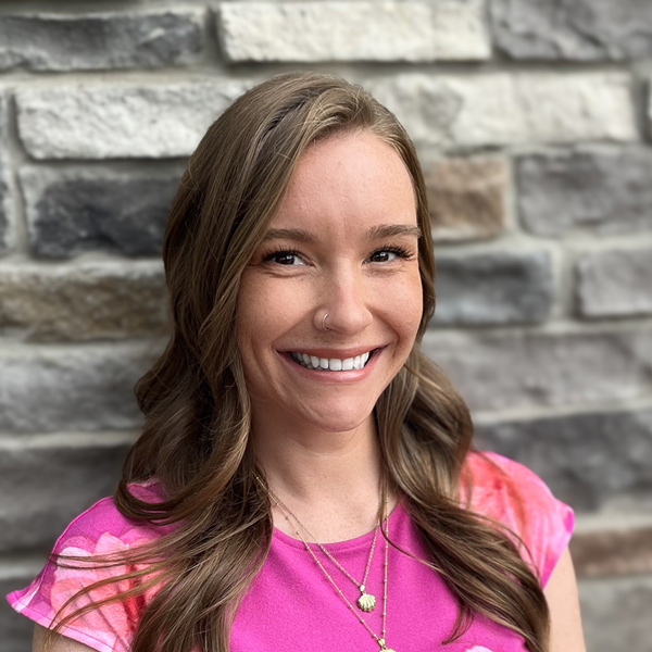 Hannah Steill, Senior Living Coordinator at Vitalia Strongsville, smiling in a professional headshot with long wavy light brown hair, layered necklaces, a bright pink top, and a gray stone wall background.
